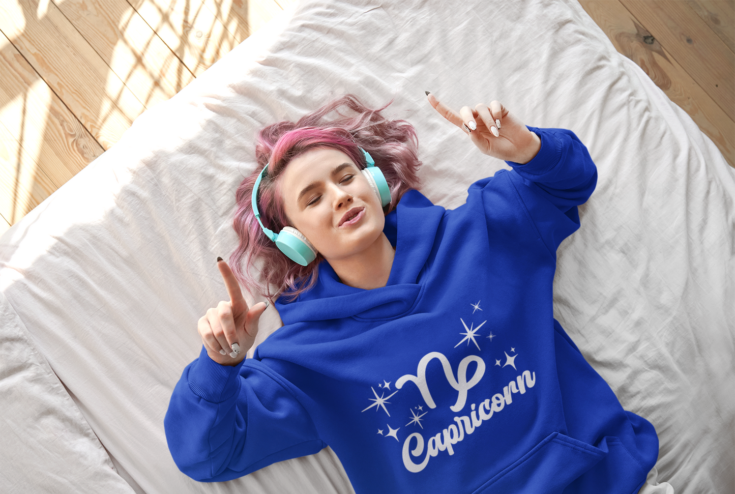 Person wearing an blue 'Capricorn' hoodie - white text design, lying on a bed with pink hair and blue headphones.