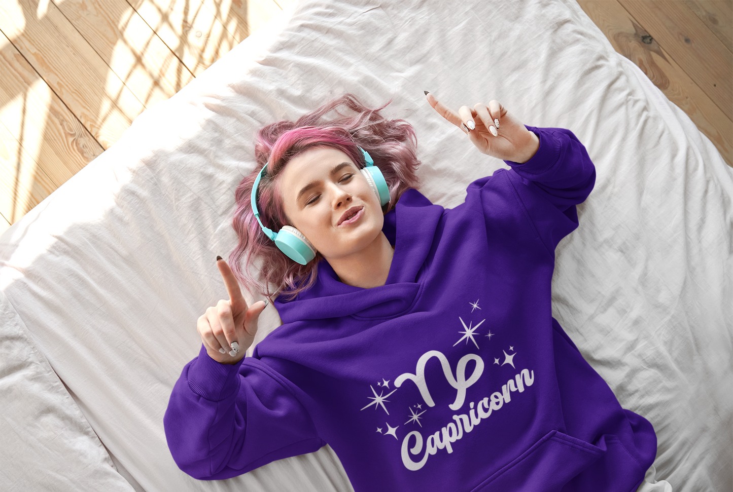 Person wearing an purple 'Capricorn' hoodie - white text design, lying on a bed with pink hair and blue headphones.