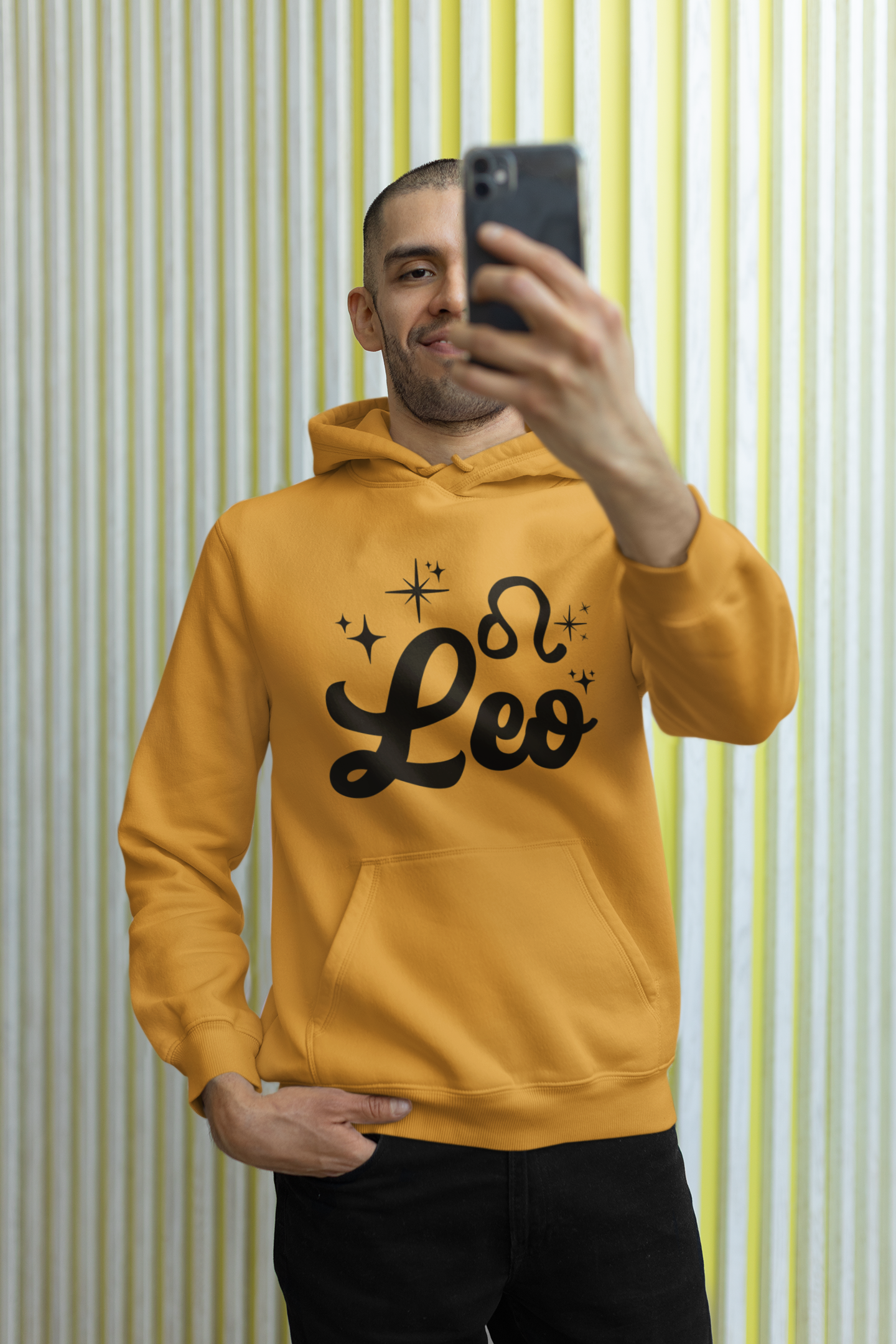 Person wearing a gold hoodie with 'Leo' black text, taking a selfie against a striped wall.