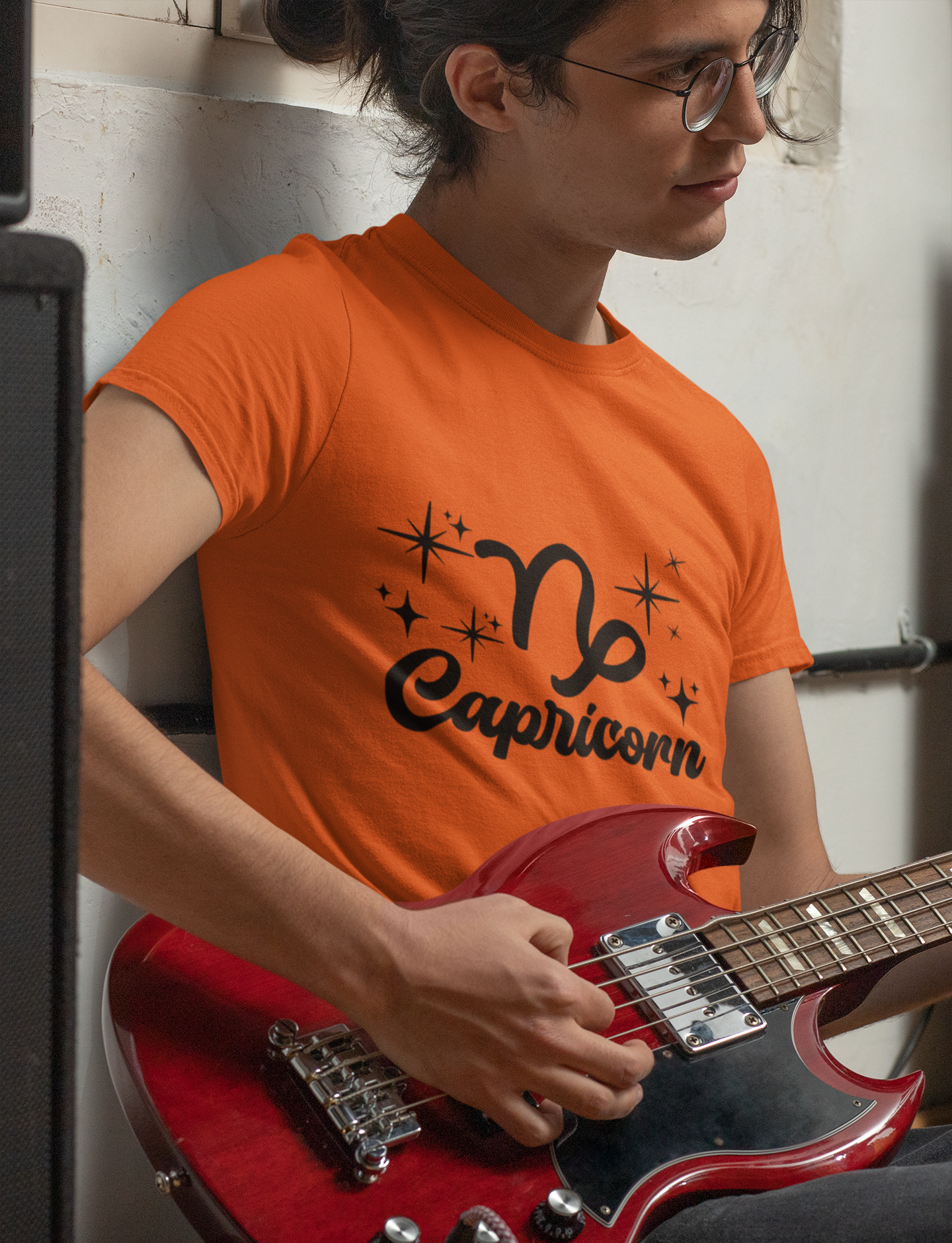 Person wearing a orange t-shirt with 'Capricorn' black text design holding a red guitar.