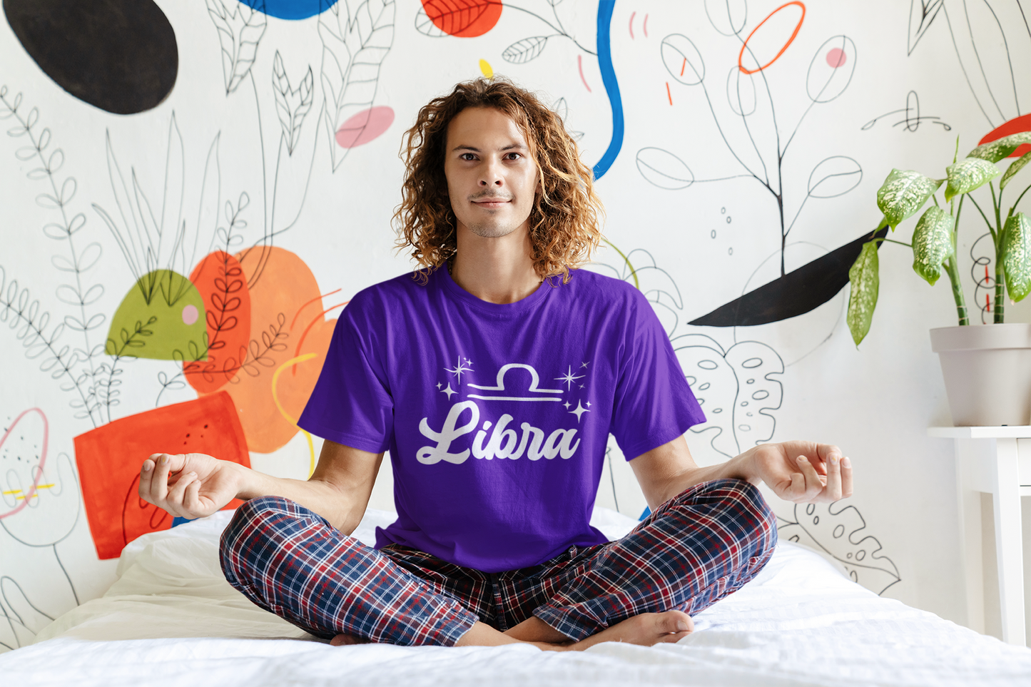 Person wearing an purple 'Libra' white text design t-shirt sitting on a bed with a colorful wall in the background