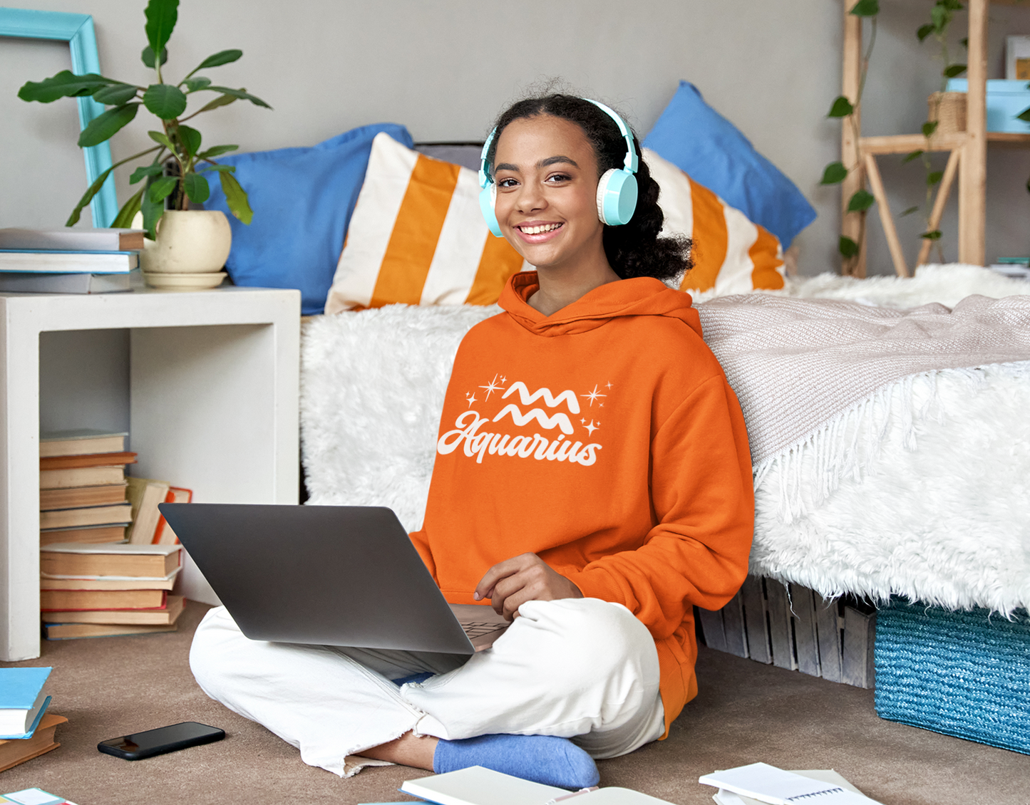 Person wearing a orange hoodie 'Aquarius' white text with zodiac symbol, sitting on a bed with a laptop, surrounded by books and a plant.