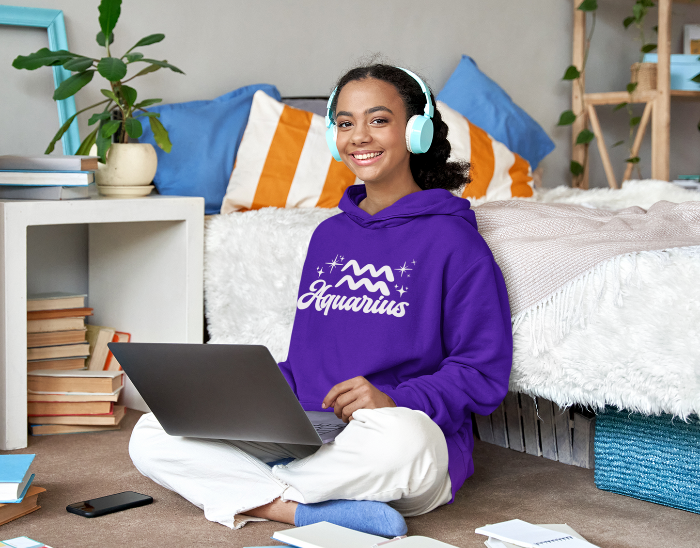 Person wearing a purple hoodie 'Aquarius' white text with zodiac symbol, sitting on a bed with a laptop, surrounded by books and a plant.
