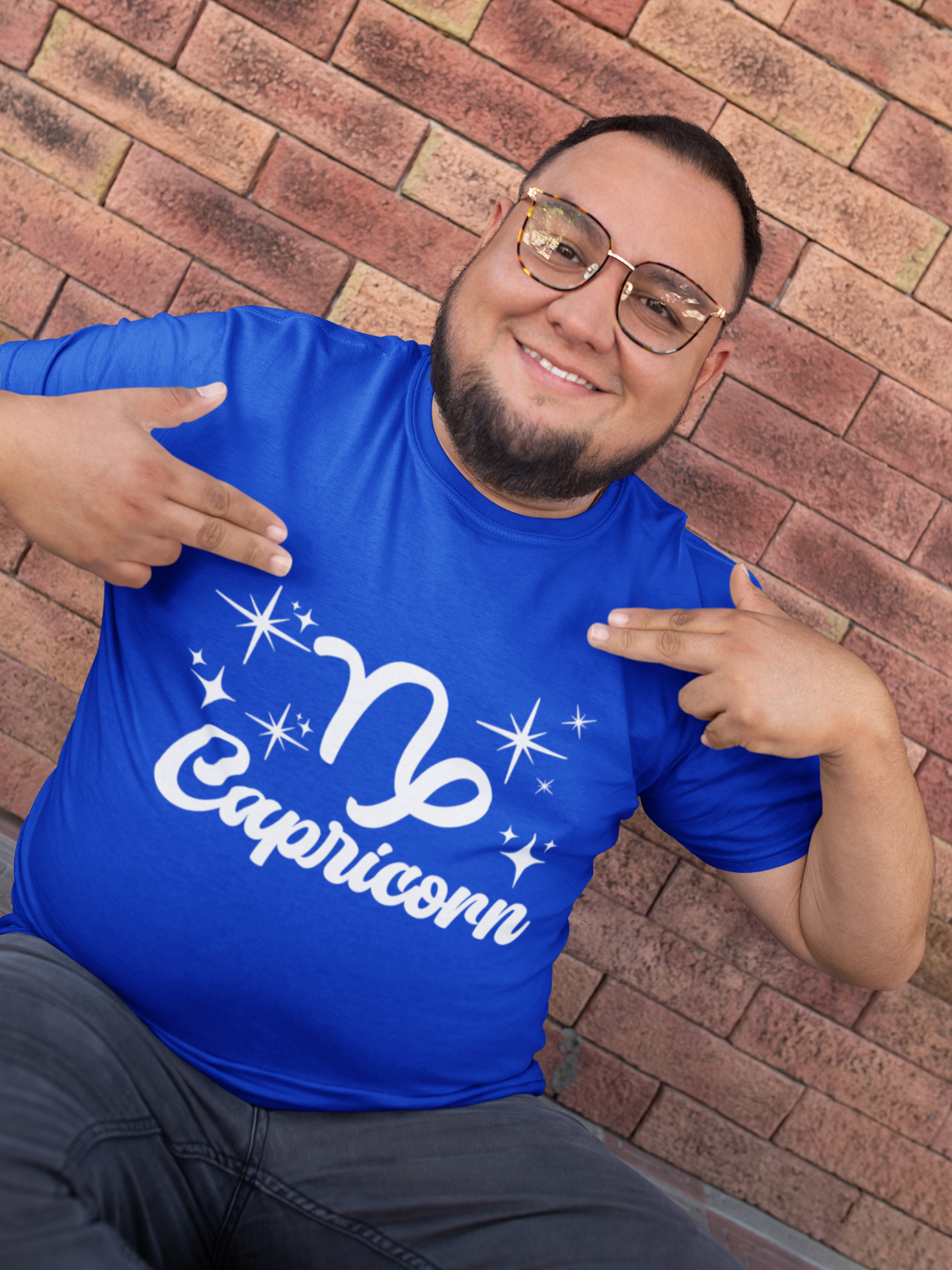 Man wearing a blue t-shirt with 'Capricorn' white text design, pointing at it, against a brick wall.