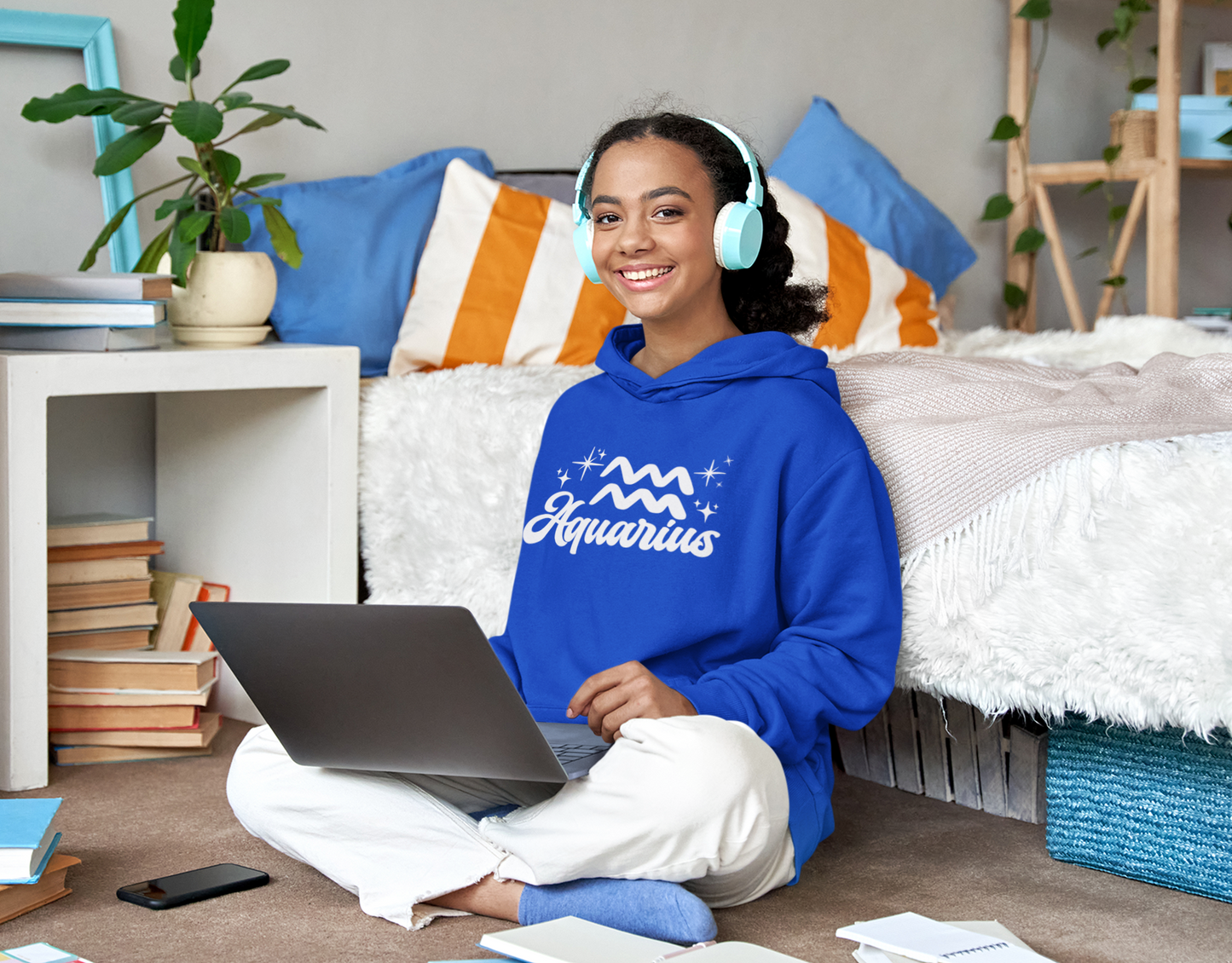 Person wearing a blue hoodie 'Aquarius' white text with zodiac symbol, sitting on a bed with a laptop, surrounded by books and a plant.