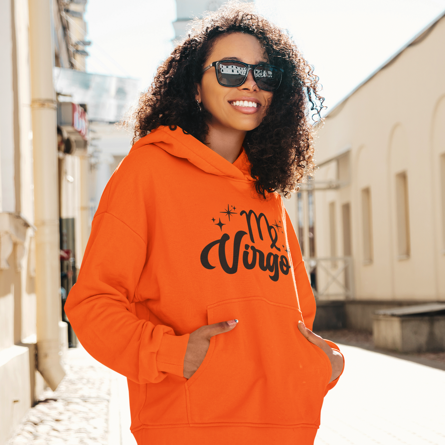 Woman with sunglasses, wearing a orange hoodie with "Virgo' black text design standing on the street.