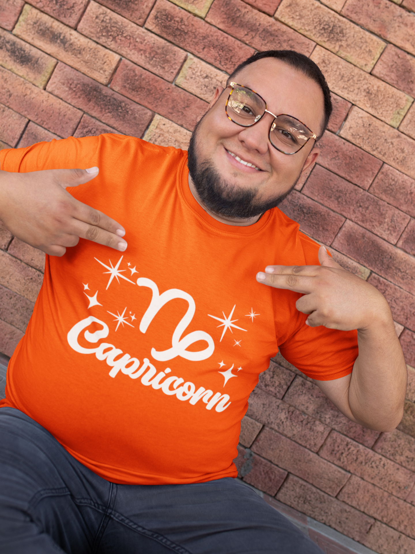 Man wearing a orange t-shirt with 'Capricorn' white text design, pointing at it, against a brick wall.