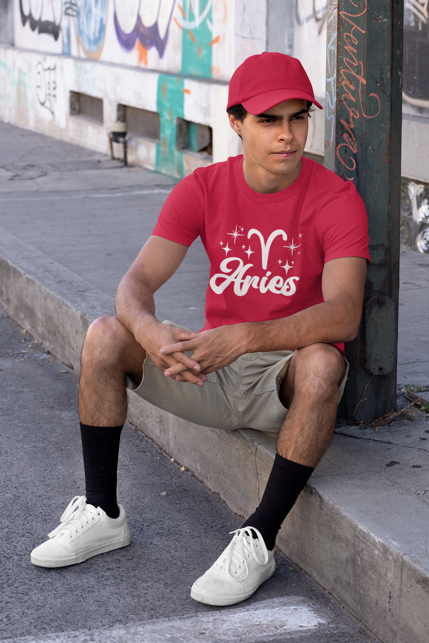 Person wearing a red 'Aries' white text design t-shirt sitting on a sidewalk.