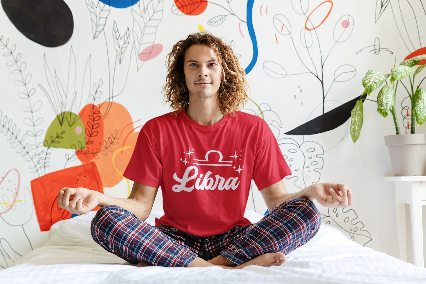 Person wearing an red 'Libra' white text design t-shirt sitting on a bed with a colorful wall in the background