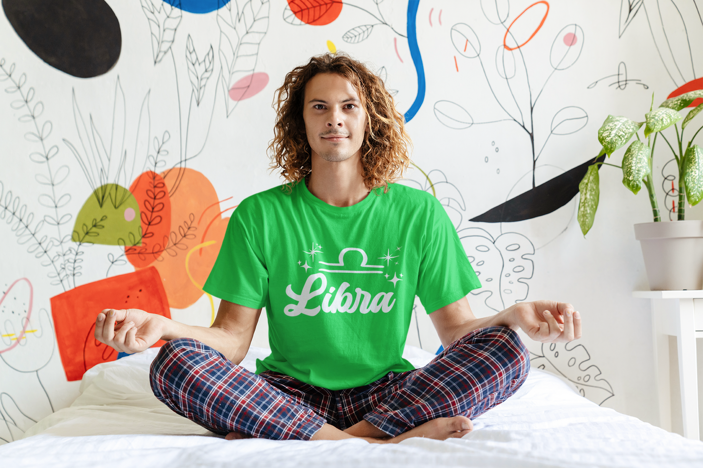 Person wearing an green 'Libra' white text design t-shirt sitting on a bed with a colorful wall in the background
