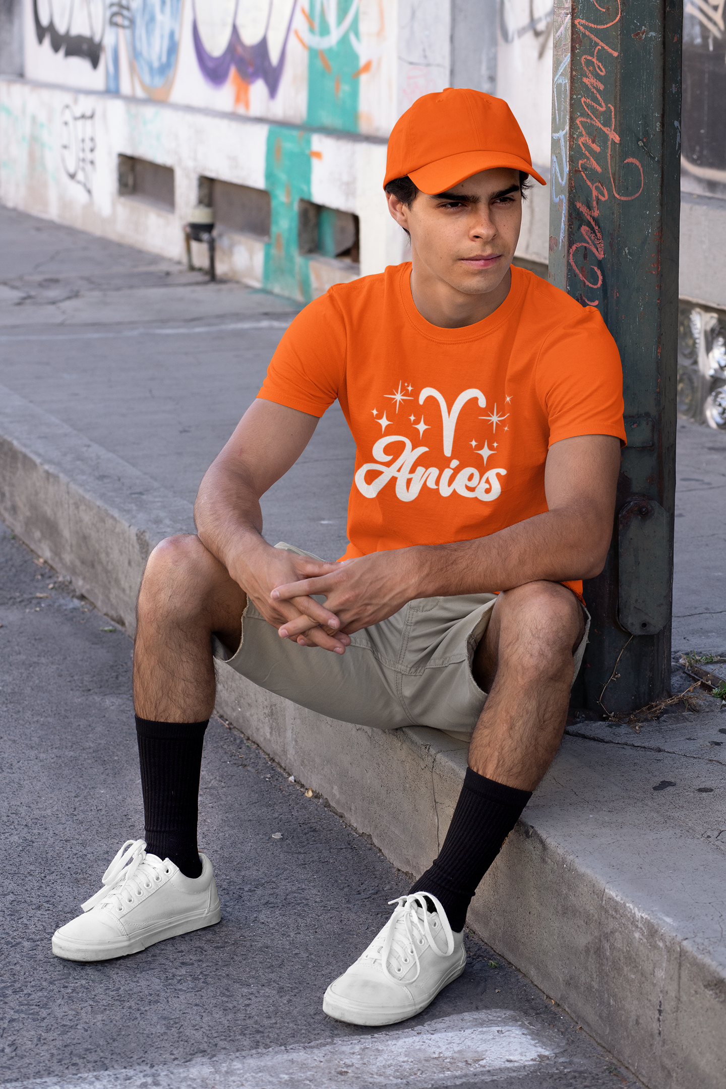 Person wearing a orange 'Aries' white text design t-shirt sitting on a sidewalk.