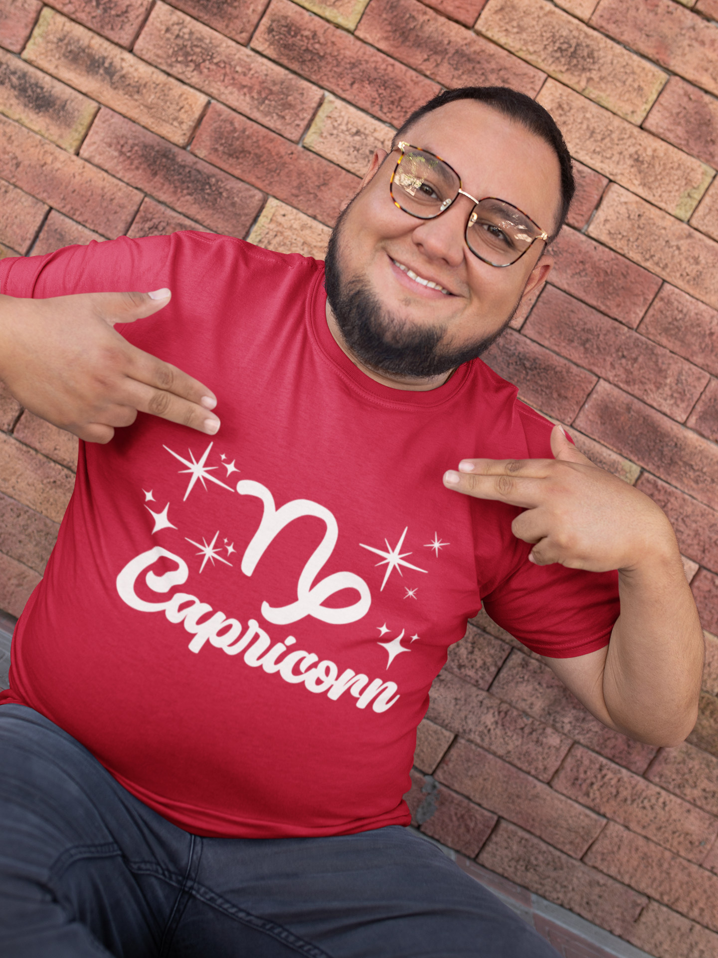 Man wearing a red t-shirt with 'Capricorn' white text design, pointing at it, against a brick wall.