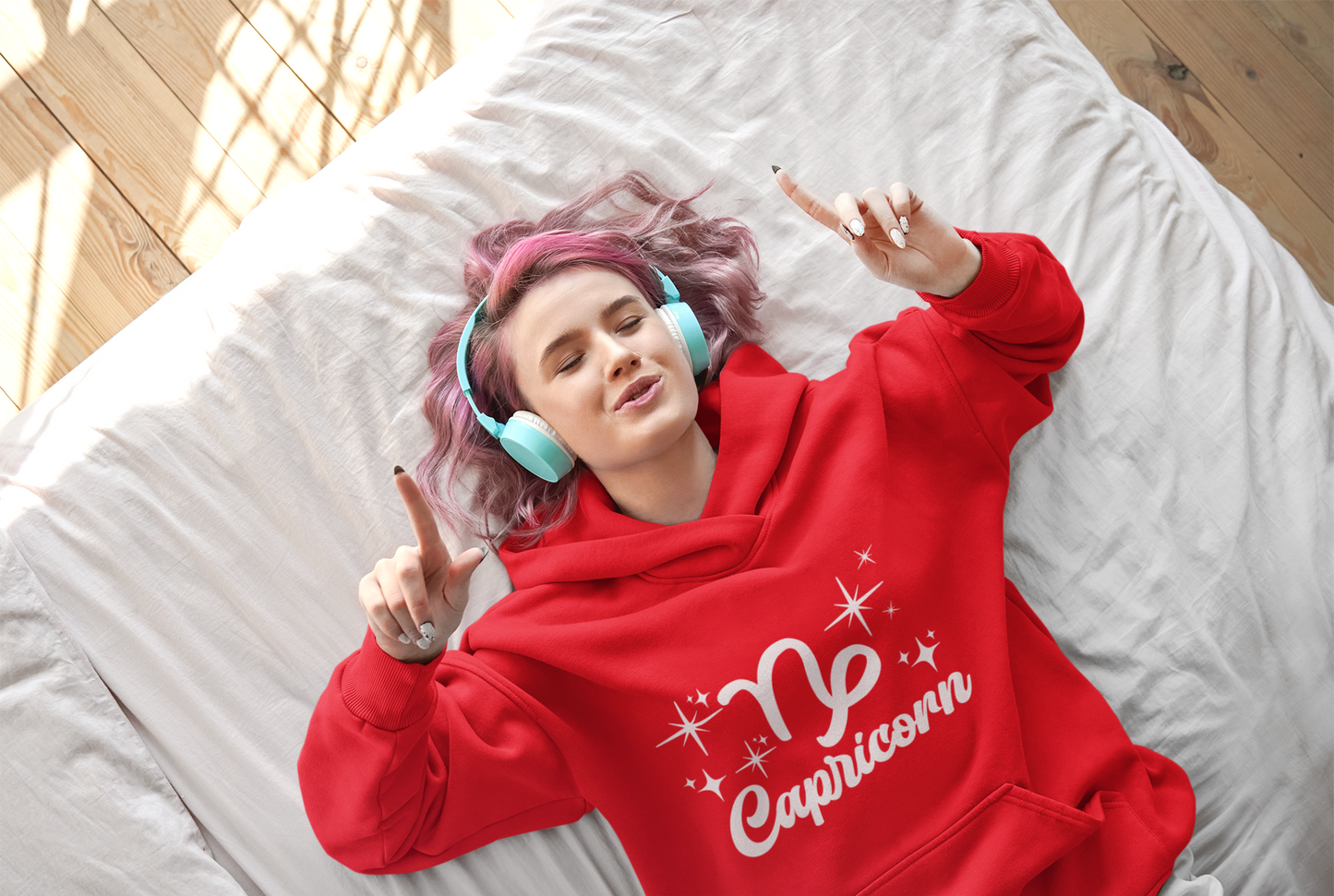 Person wearing an red 'Capricorn' hoodie - white text design, lying on a bed with pink hair and blue headphones.