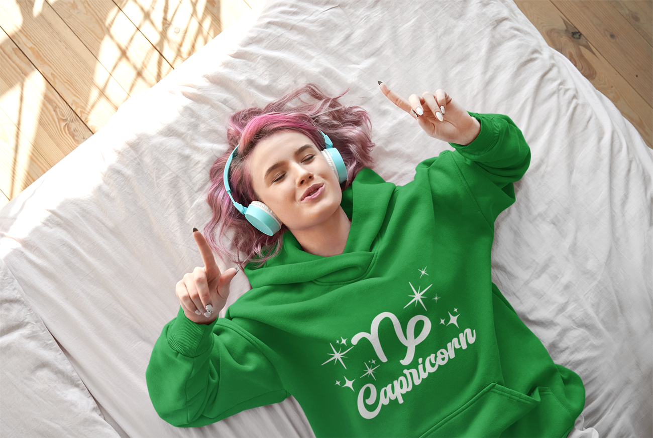 Person wearing an green 'Capricorn' hoodie - white text design, lying on a bed with pink hair and blue headphones.