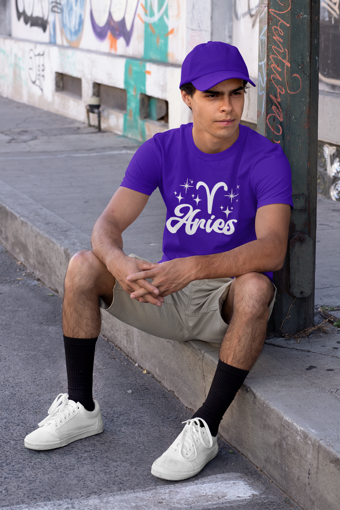 Person wearing a purple 'Aries' white text design t-shirt sitting on a sidewalk.