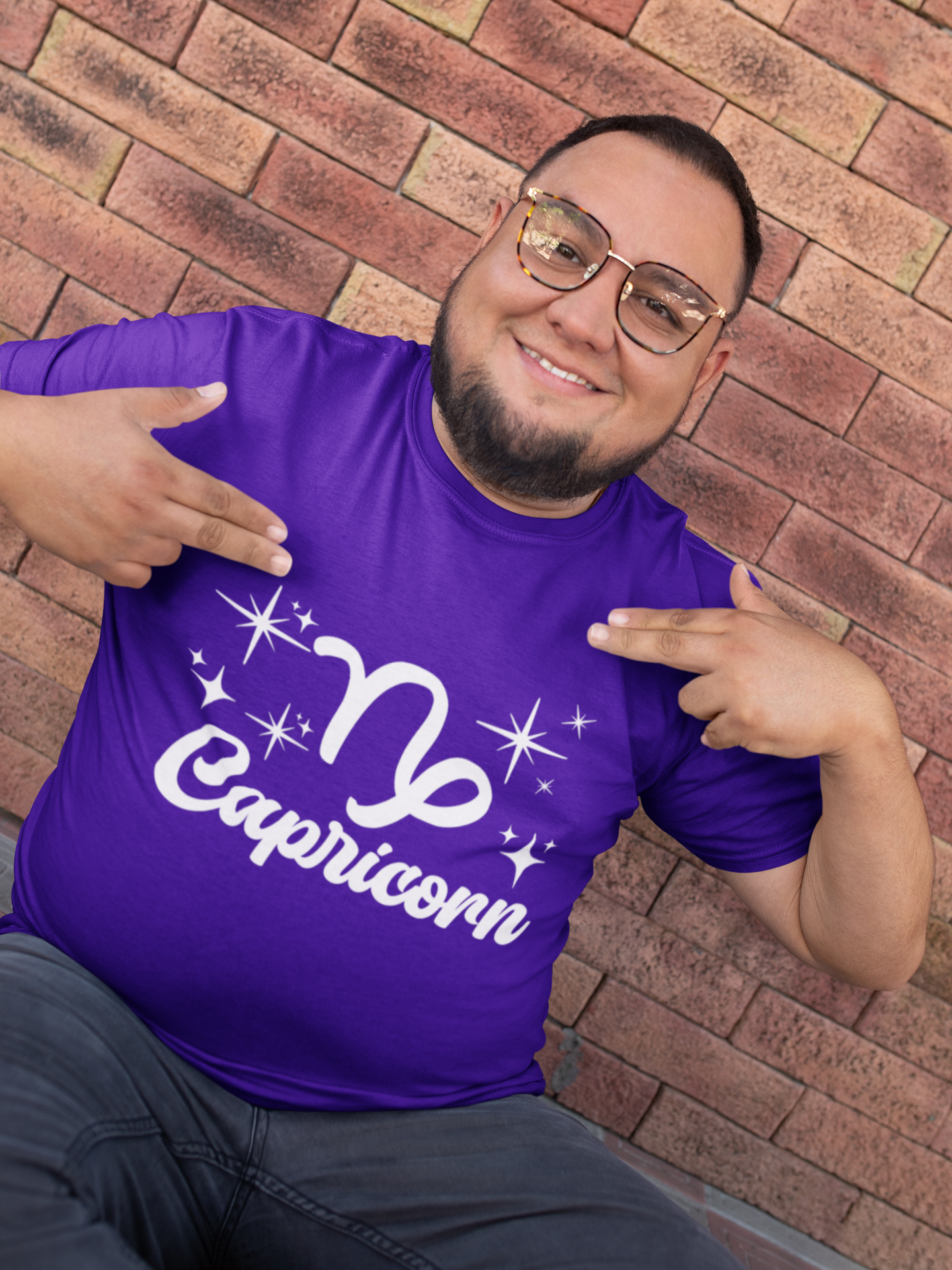 Man wearing a purple t-shirt with 'Capricorn' white text design, pointing at it, against a brick wall.