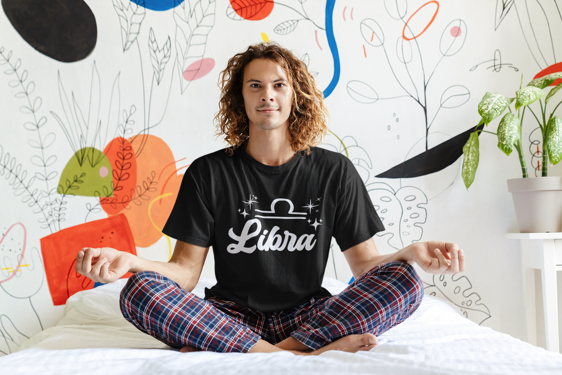 Person wearing an black 'Libra' white text design t-shirt sitting on a bed with a colorful wall in the background
