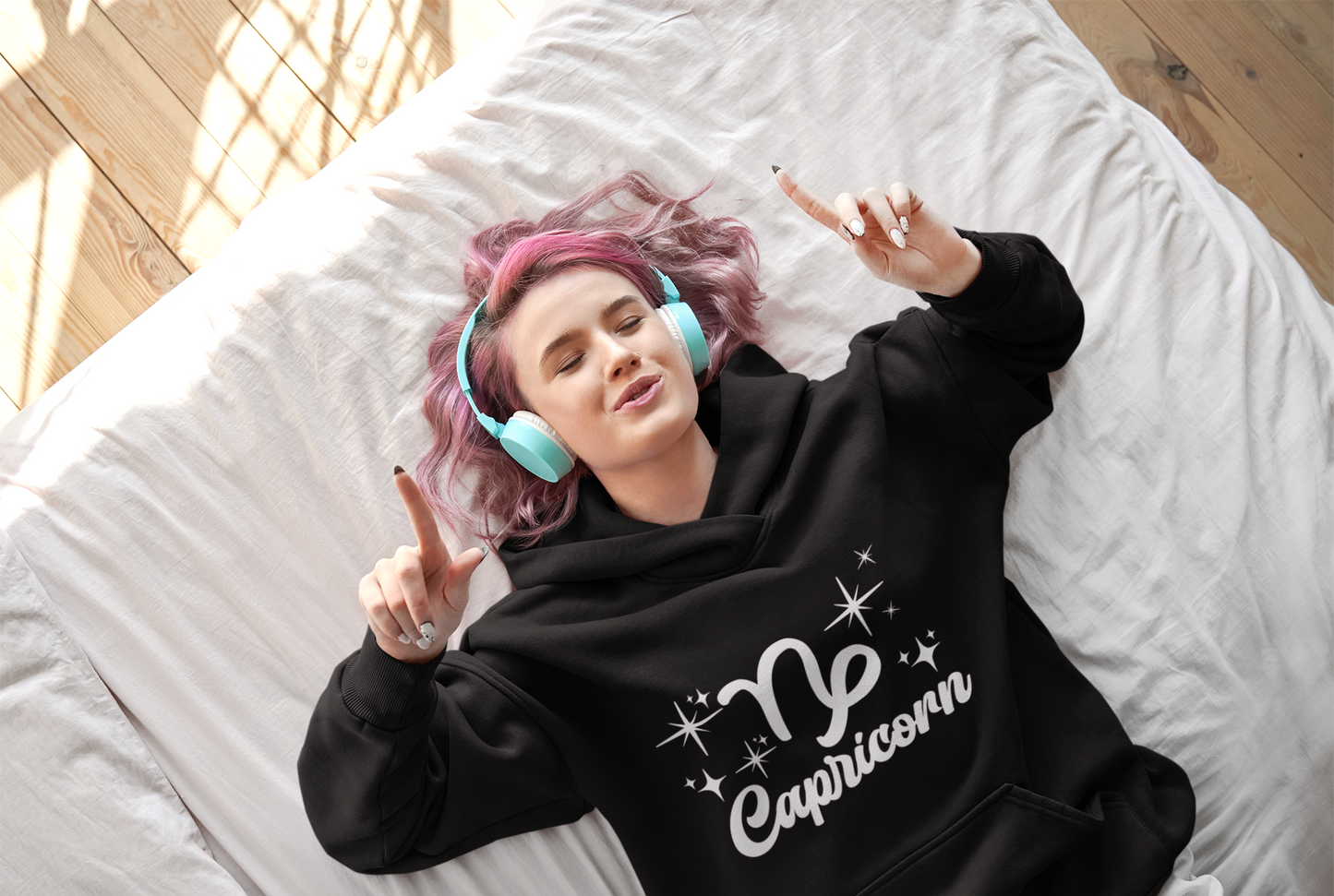Person wearing an black 'Capricorn' hoodie - white text design, lying on a bed with pink hair and blue headphones.
