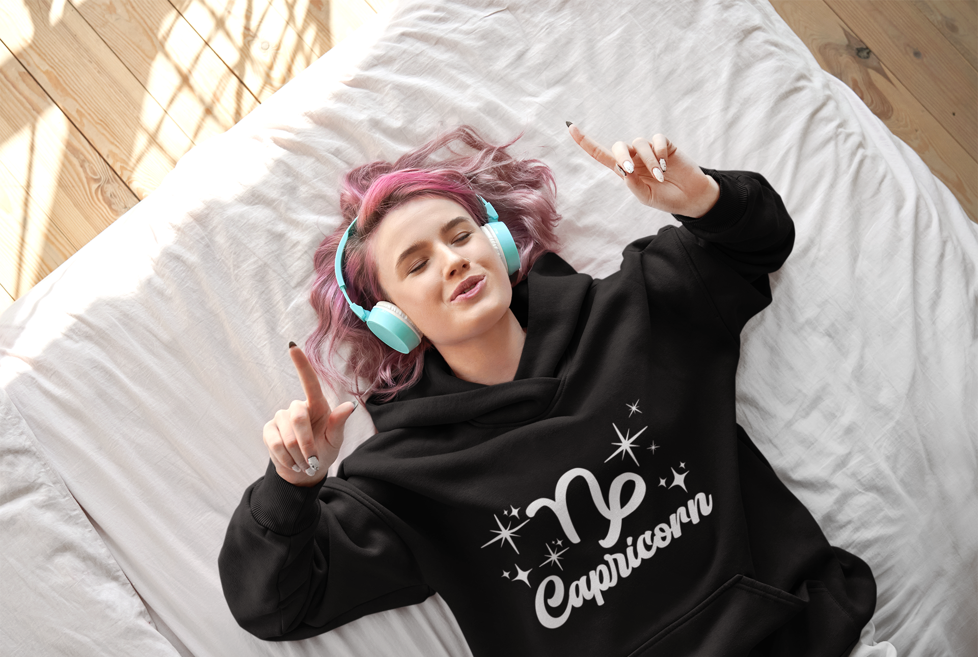 Person wearing an black 'Capricorn' hoodie - white text design, lying on a bed with pink hair and blue headphones.