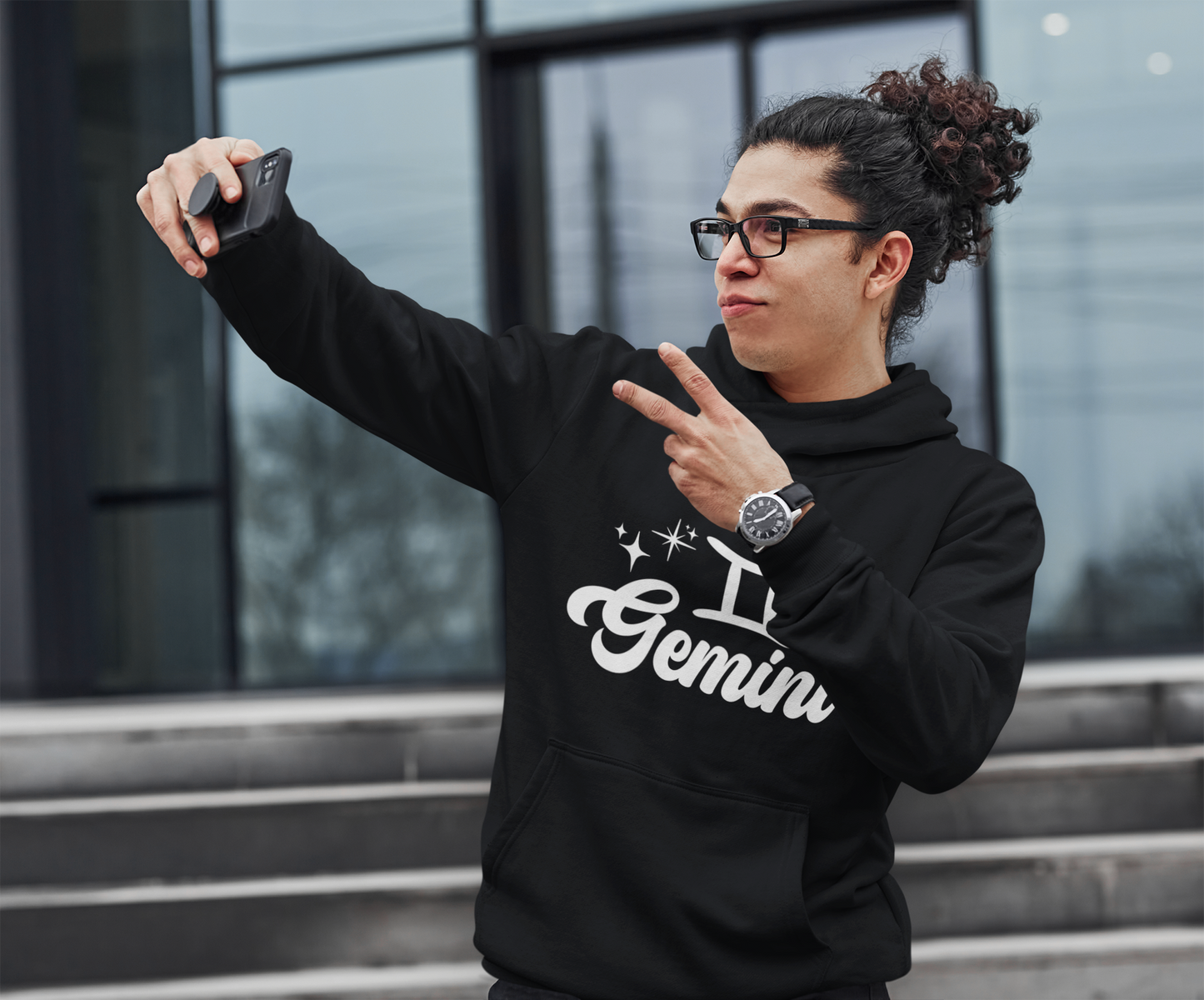Person wearing an black hoodie with 'Gemini' white text, taking a selfie outdoors.