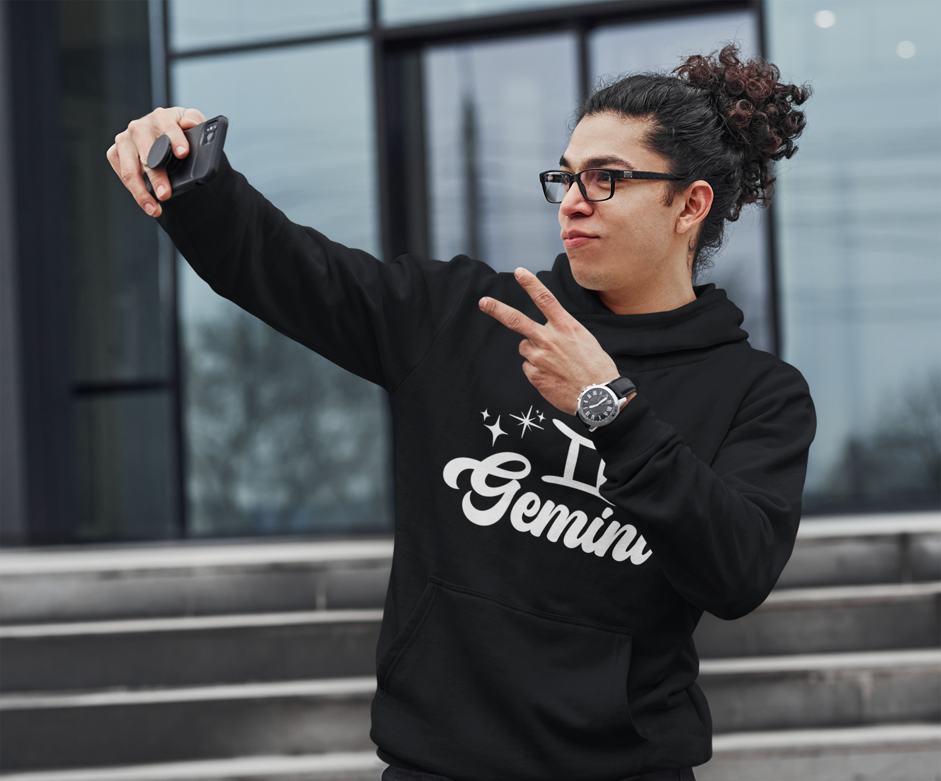 Person wearing an black hoodie with 'Gemini' white text, taking a selfie outdoors.