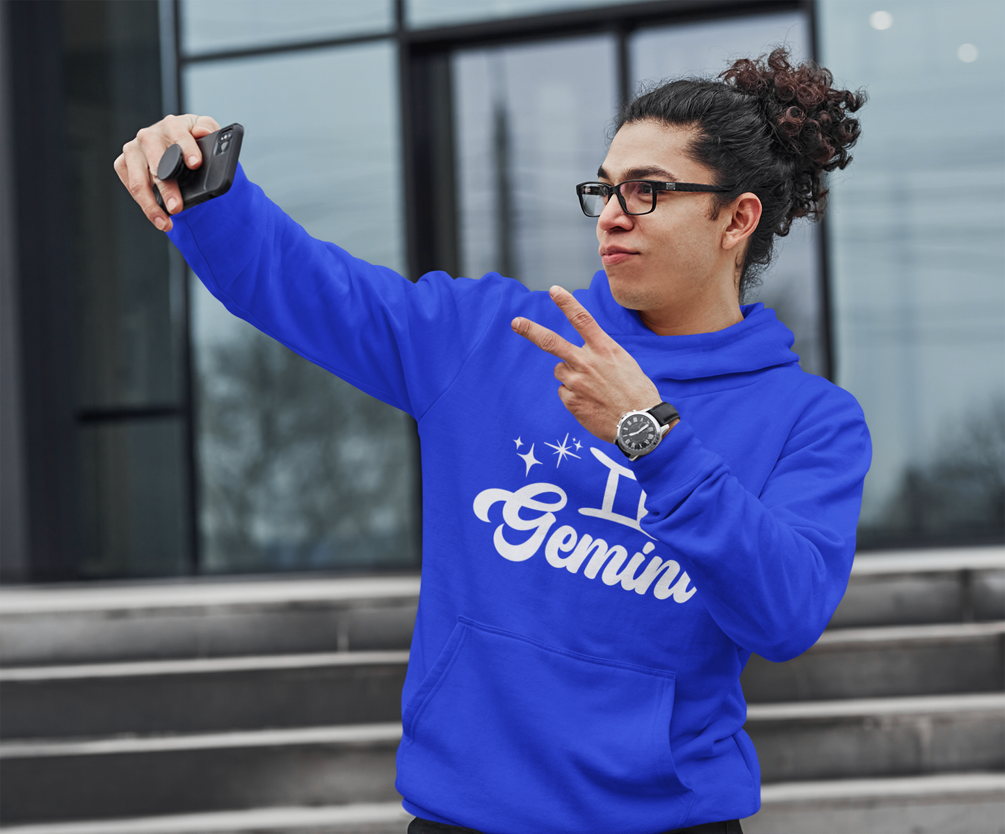 Person wearing an blue hoodie with 'Gemini' white text, taking a selfie outdoors.