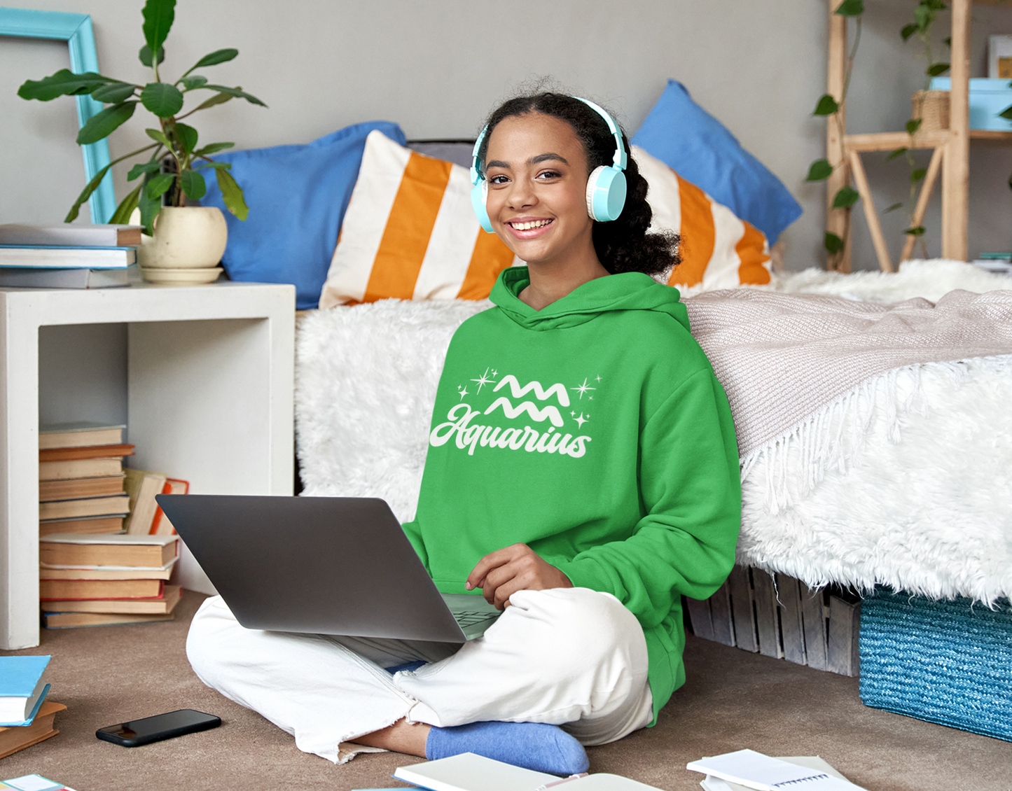 Person wearing a green hoodie 'Aquarius' white text with zodiac symbol, sitting on a bed with a laptop, surrounded by books and a plant.