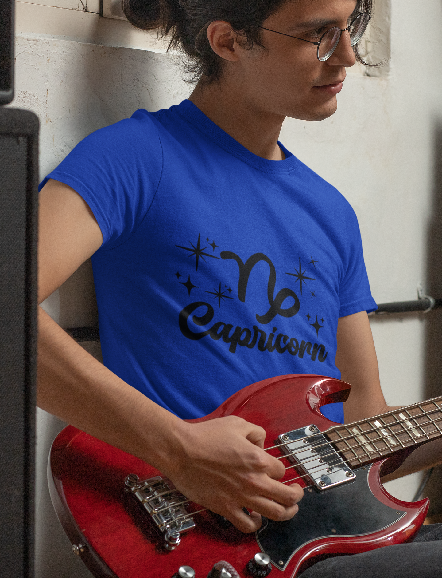 Person wearing a blue t-shirt with 'Capricorn' black text design holding a red guitar.